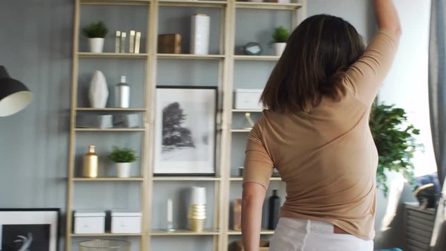 Attractive stylish caucasian woman in glasses in her twenties dances enthusiastically summersault enjoys herself alone and sings moving around the grey room in scandinavian style with golden bookcase