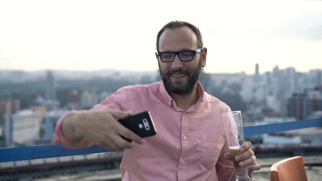 Young man with drink taking selfie with cellphone in rooftop bar
