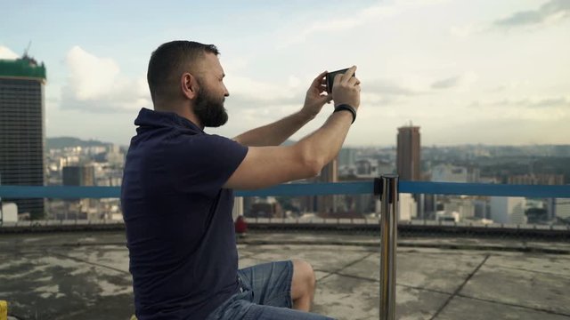 Young man taking photos of cityscape view with cellphone in rooftop bar
