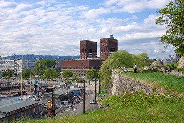 Widk centrum Oslo, stolicy Norwegii, z fortecznego muru, na którym ludzie siedzą, stoją, relaksują się, charakterystyczne budowle miasta, zatoka morska z jachtami, spacerującymi ludźmi - obrazy, fototapety, plakaty