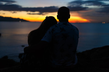 Silhouette of a couple are sitting and hugging on the beach at sunrise. Back view