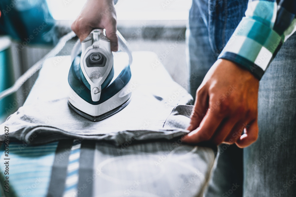 Wall mural Man at home ironing clothes. Father performs home duties, irons the child's clothes. Man in role of a woman at home. Man ironing shirt on ironing board with steaming blue iron.