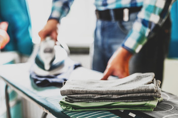 Man at home ironing clothes. Father performs home duties, irons the child's clothes. Man in role of a woman at home. Man ironing shirt on ironing board with steaming blue iron.