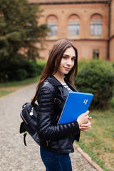 Obraz premium Portrait of an attractive smiling student girl standing with backpack and books and looking away outdoors