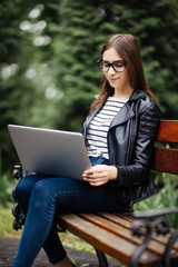 Beautiful young woman sitting in public park chatting over laptop computer. Young student in campus park.