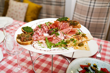 Meat antipasto platter on dinner table. Cold meat plate with prosciutto, salami, ham, bacon, sandwiches and olives on white plate, copy space. Italian prosciutto crudo or jamon with arugula. Raw ham
