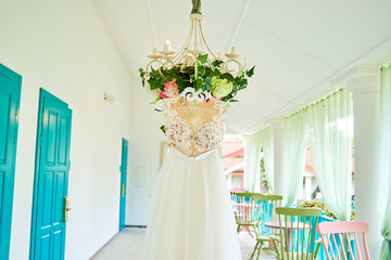 Beautiful long wedding dress for bride hanging on hanger with inscription in Russian ''Wedding'' on chandelier with flowers on hotel terrace. Wedding fashion, copy space