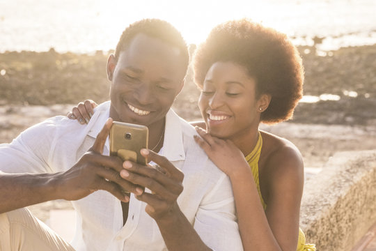 Happy Black Young Couple At The Beach With Sunset Backlight On The Background, Smile And Laugh Using A Smartphone To Chat With Friends Or To See Pictures Of The Vacation