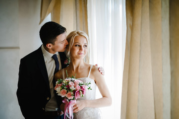 Stylish bride with a wedding bouquet of flowers and groom near the window indoors in the rooms. Man...