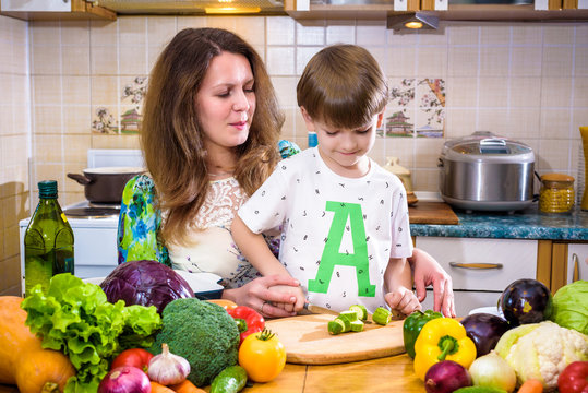 The young cook mother standing with her little son in the kitchen and salting vegetables