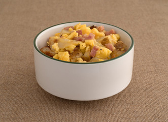 A bowl filled with scrambled eggs with potatoes cheese and ham on a brown tablecloth.