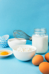 Eggs of chicken, flour in a bowl, sugar, milk in a glass jar whisk ingredient for cooking a bake on a blue background with copy space