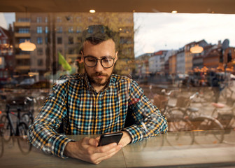 fashion guy with glasses and checkered shirt sitting in a cafe working in the phone