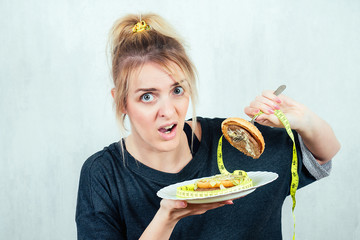 portrait of a hungry, young and beautiful blonde woman holding a burger (cheeseburger) on a plate in a black T-shirt and measuring tape. concept of diet and rejection of unhealthy harmful food