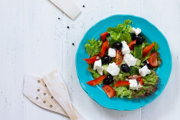 Greek salad with fresh vegetables, feta cheese and black olives in a blue plate on white wooden table. Copy space, top view flat lay background.