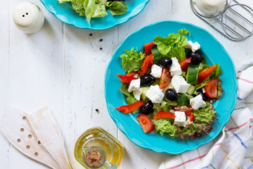 Greek salad with fresh vegetables, feta cheese and black olives in a blue plate on white wooden table. Top view flat lay background.