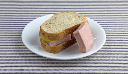 Pressed pork canned meat sandwich on wheat bread with a chunk to the side on a white plate atop a blue striped tablecloth.