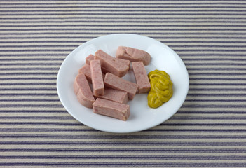 Pressed pork canned meat chunks on a plate with mustard on a blue striped tablecloth.