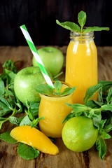 Yellow smoothie in glass jars surrounded by fruits and mint against wooden background