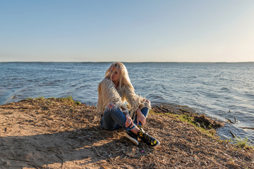 sad grim blond girl sitting on the seashore with tousled hair in lifestyle clothes