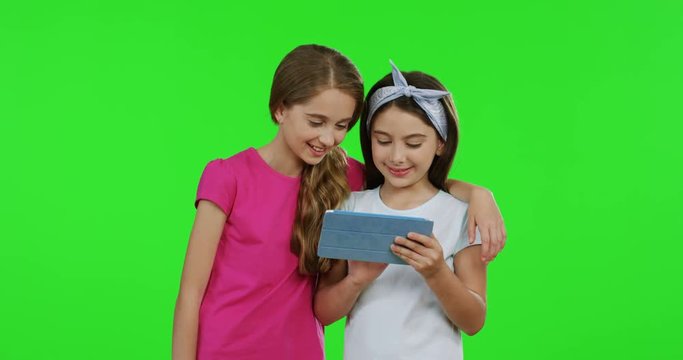 Two lovely teenage friends standing on the green screen background and scrolling on the tablet computer screen. Chroma key