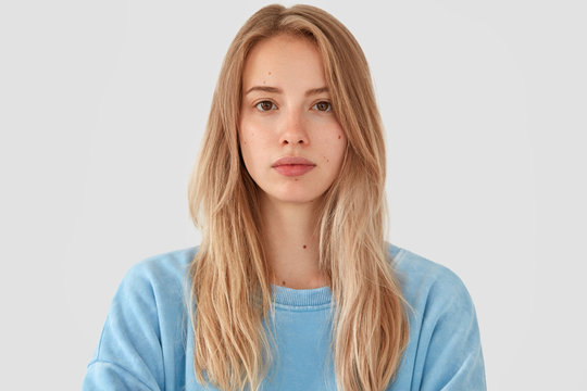 Headshot of tender beautiful young female model with fair hair, wears blue top, has healthy pure skin, looks seriously at camera, poses against white concrete wall. Cute teenager feels confident