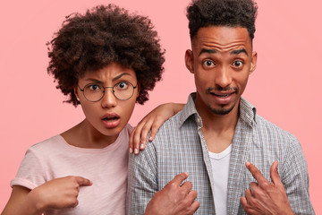 Puzzled dark skinned woman and man look in bewilderment and frustration at camera, being shocked to be chosen, indicate at themselves. Surprised young African American couple gesture indoor.