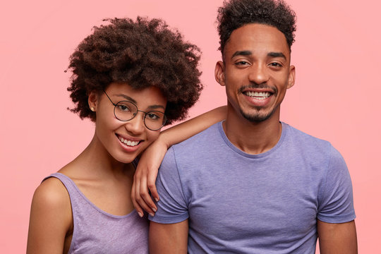 Positive dark skinned female with Afro hairstyle leans at shoulder of her best mixed race male friend, enjoy spending time together, pose against pink background. African couple: wife and husband