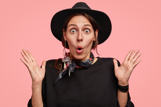 Emotional female adult gestures with stunned expression, stares at camera, wears black elegant hat, being surprised by unexpected gift, poses against pink background. People and emotions concept