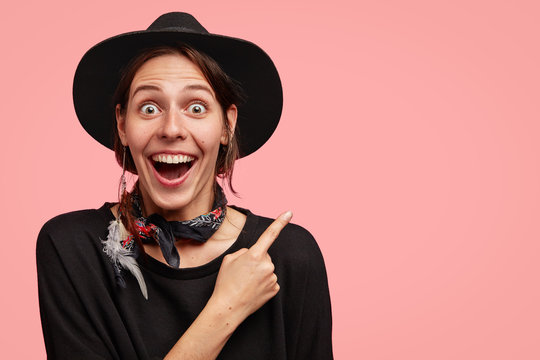 Horizontal portrait of overjoyed beautiful female wears cowboy style of clothes, indicates at upper right corner with fore finger, stares at camera, isolated over pink background, advertises something