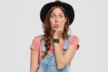 Portrait of adorable astonished female model with surprised expression, recieves unexpected offer, keeps hands on cheek and gasps from shock, wears black hat, stands indoor. Omg, what I see!