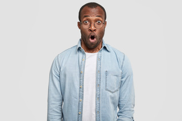 I don`t believe it`s real true! Portrait of excited unshaven African American male keeps mouth widely opened, stares at camera with unexpectedness, poses on white background. Stunned dark skinned guy