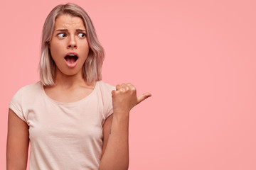 Studio shot of amazed stupefied female model looks with amazement aside, indicates at blank copy space, isolated on pink background. Thrilled adorable student with stylish haircut gestures indoor