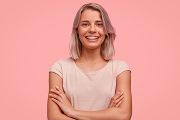 Horizontal shot of pretty successful young female blogger, keeps hands crossed, glad to achieve success, has dyed hair, wears braces on teeth and casual clothing, isolated on pink background