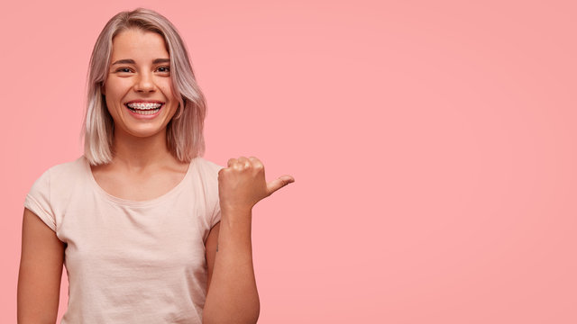 This is perfect place for entertaining. Positive good looking female model with bobbed hairstyle, indicates with thumb at blank copy space, raises hand, blank pink wall for your advertisement