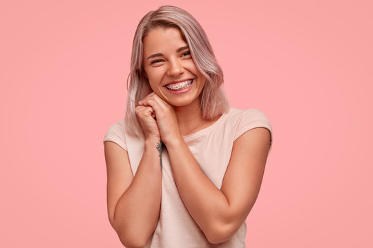 Carefree beautiful female with braces on teeth, smiles broadly at camera, being in high spirit while hangs out with friends, has joyful expression, stands against pink background. Optimistic girl