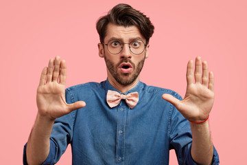 Surprised bearded male with trendy hairdo, has surprised expression, stretches hands as shows stop gesture, refusing organising party, wears elegant stylish denim shirt and spectacles, isolated