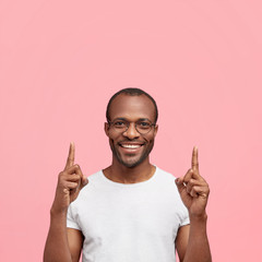Positive African American male in casual outfit, smiles broadly from satisfaction, points with fore...