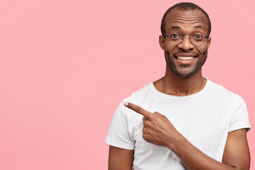 Glad unshaven dark skinned male with positive expression, wears casual white t shirt and spectacles, indicates at blank copy space with fore finger, happy to advertise something. Horizontal shot