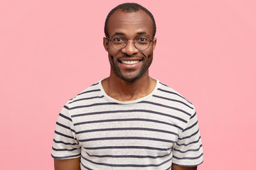Handsome delighted guy with dark skin, pleasant smile, wears casual striped t shirt, poses against...