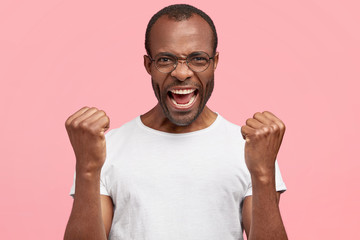 Emotional annoyed unshaven dark skinned young male clenches fists in anger, wears casual white t...