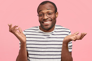 Happy hesitant African American male with cheerful expression, shrugs shoulders in bewilderment,...