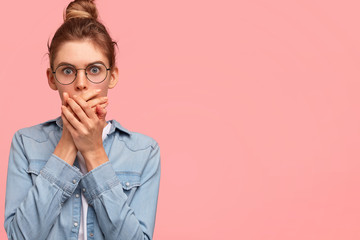 Portrait of attractive female being shocked with prices during shopping, covers mouth with palms, expresses great surprisement, wears denim jacket, stands against pink background, blank space