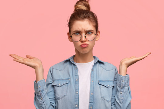 Portrait of puzzled intelligent female student wears big spectacles, feels awkward, shrugs shoulders with hesitation, sees no way out, doesn`t know material on exam isolated on pink background.