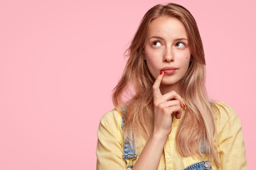Thoughtful female youngster keeps fore finger near lips, being deep in thoughts, thinks about...