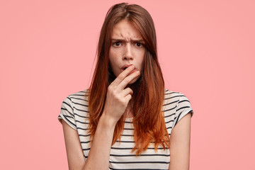 Portrait of puzzled Caucasian female with freckled skin frowns face, being displeased, has grumpy expression, wears casual striped t shirt, poses against pink background. Serious gloomy woman indoor