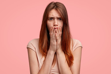 Stunned female with freckled skin, covers mouth and frowns face, has long hair, wears casual clothes, poses against pink background. Dissatisfied young woman recieves awful news from close person