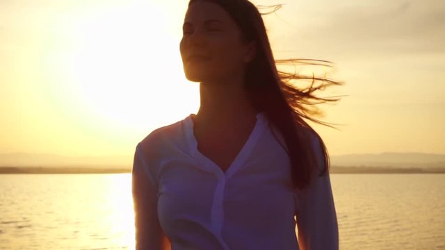 Dark silhouette of smiling young woman at sunset on lake enjoying quiet moment in nature. Close up of carefree female figure meditating at golden hour in slow motion