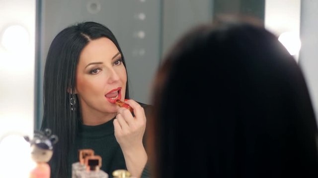 Reflection of lady applying her make-up. Brunette mature woman putting lipstick while looking in mirror. Makeup at night getting ready before going to party. Slow motion