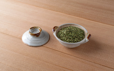Cilantro flakes in a small bowl with a lid to the side on a wood table.